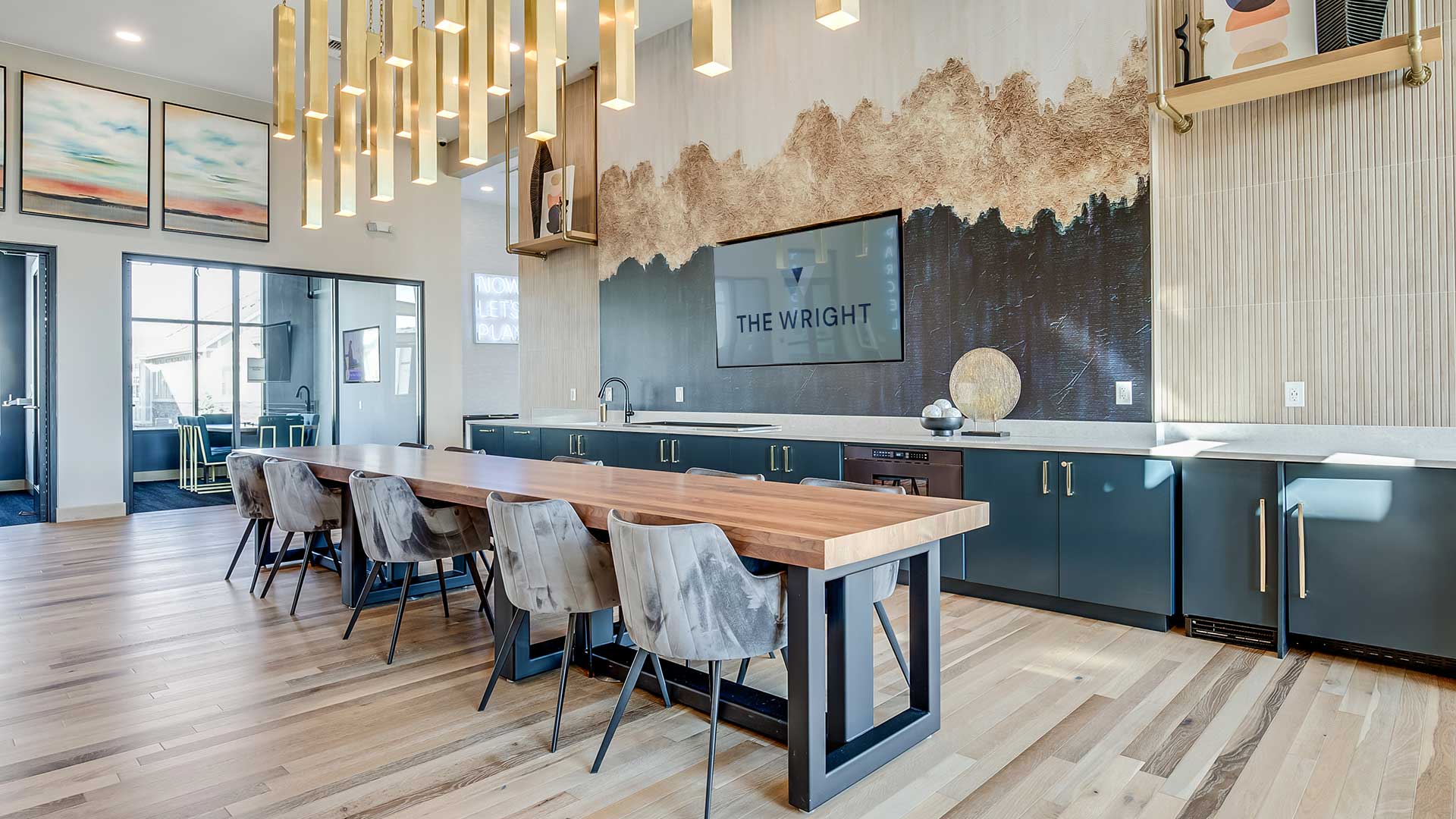 Upscale apartment clubhouse lounge with large communal dining table, modern pendant lighting, entertainment wall with mounted TV, and contemporary seating areas.