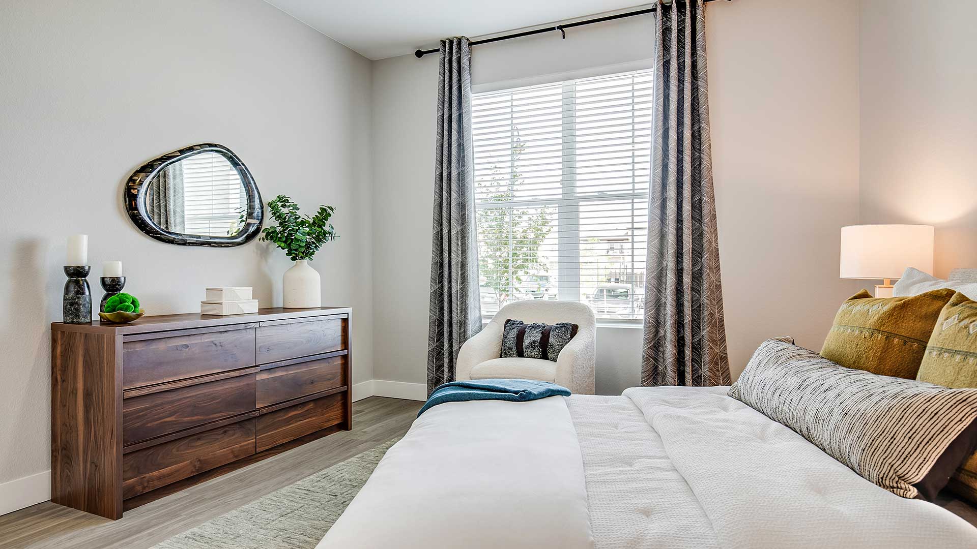 Modern apartment bedroom with large window, wood dresser, accent chair, and neutral contemporary decor with natural light.